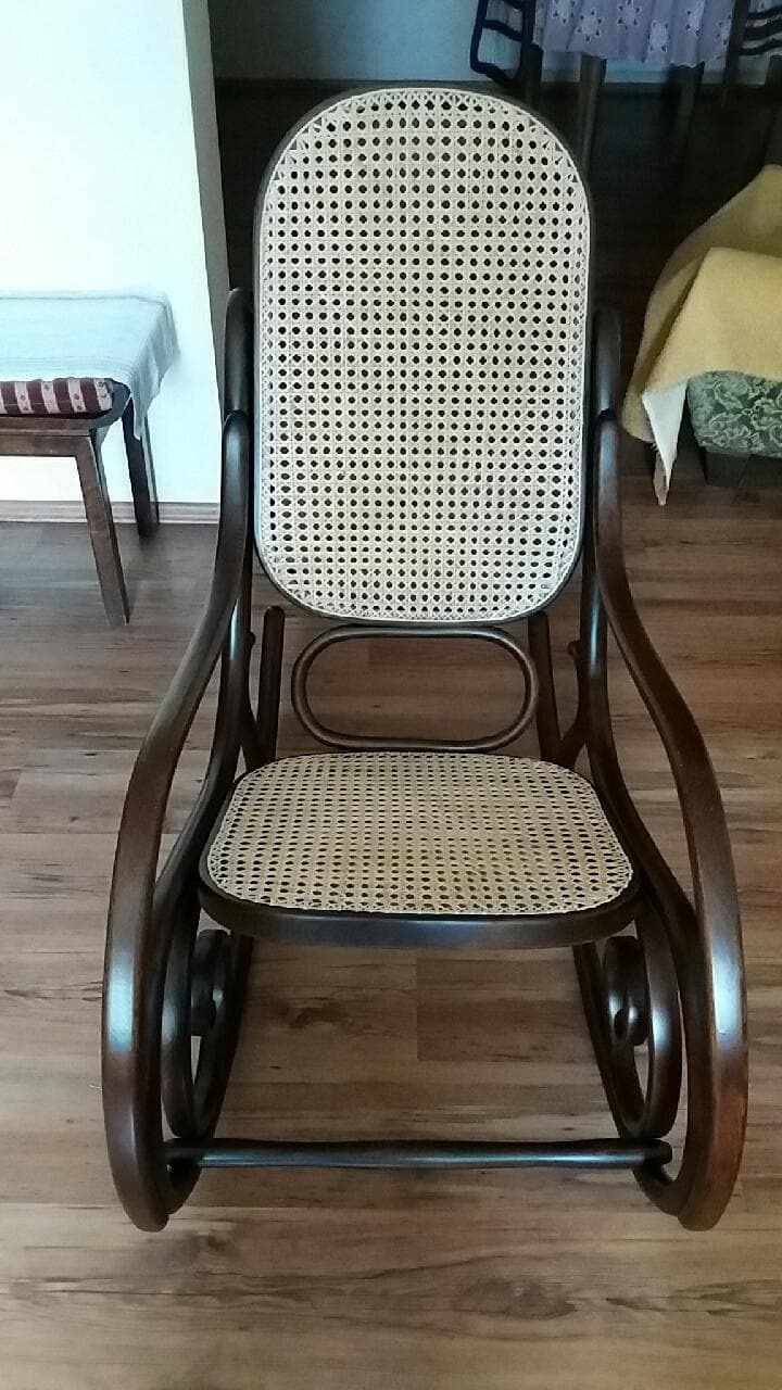 Ciemnobrązowy fotel bujany z jasną, plecioną rattanową siatką na oparciu i siedzisku, stojący na drewnianej podłodze.
