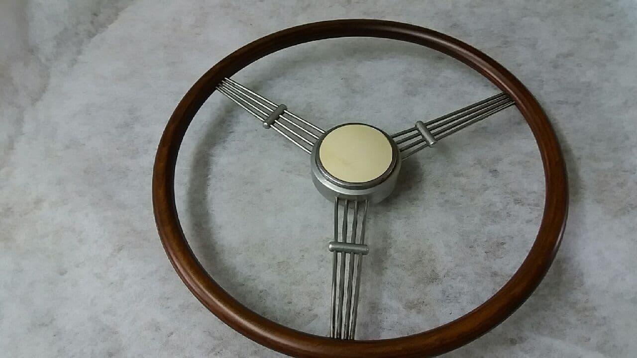 Klasyczna kierownica samochodowa z drewnianym wieńcem, metalowymi szprychami i kremowym środkiem, leżąca na szarym tle.