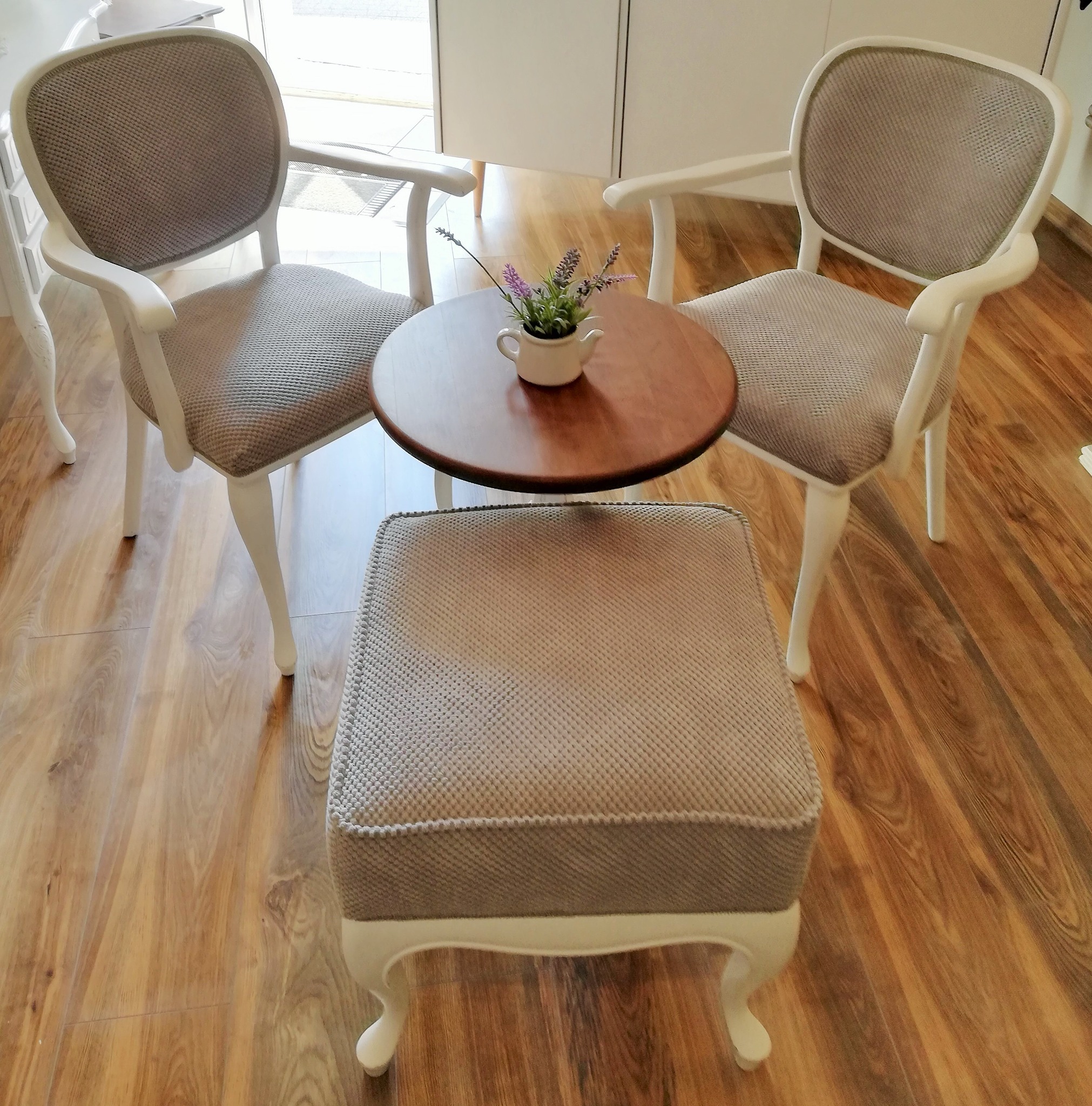 Komplet wypoczynkowy: dwa krzesła z jasną tapicerką i białymi nogami, okrągły stolik z ciemnym blatem i mały, tapicerowany puf na tle drewnianej podłogi.