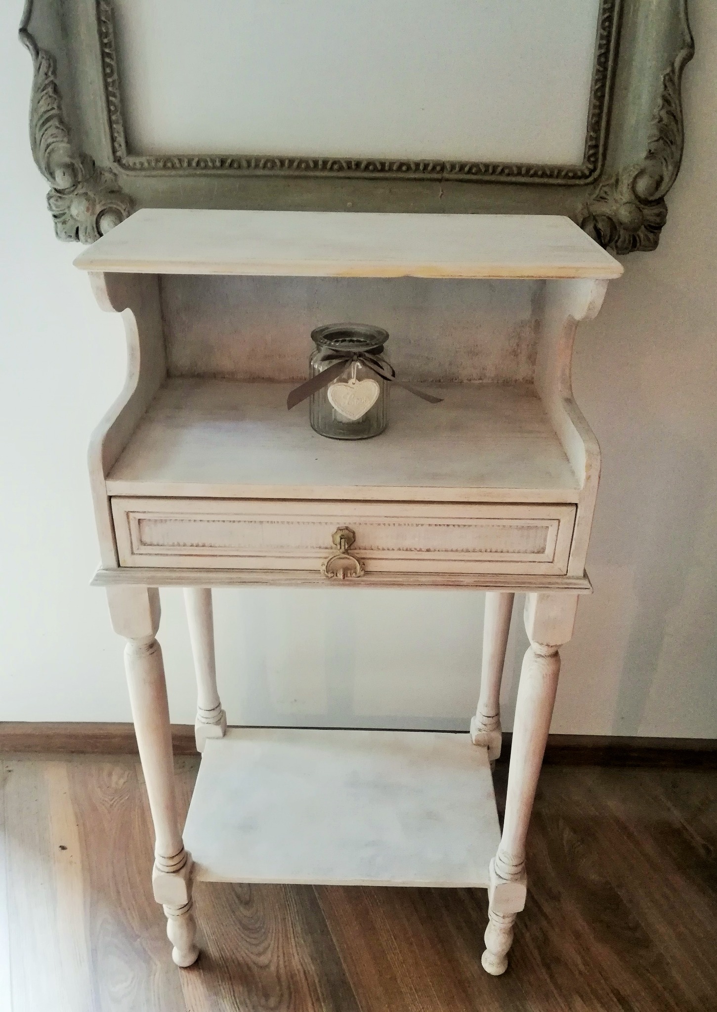 Biała, odrestaurowana szafka nocna w stylu vintage z szufladą i półką, na której stoi szklany słoik z ozdobnym sercem, w tle fragment ramy obrazu.