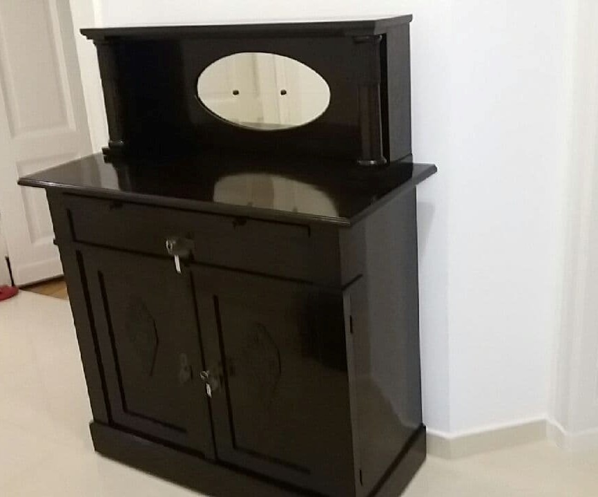 Ciemnobrązowa, lakierowana komoda z dwoma drzwiczkami, szufladą i nadstawką z owalnym lustrem, widoczny klucz w zamku szuflady, ustawiona przy białej ścianie.