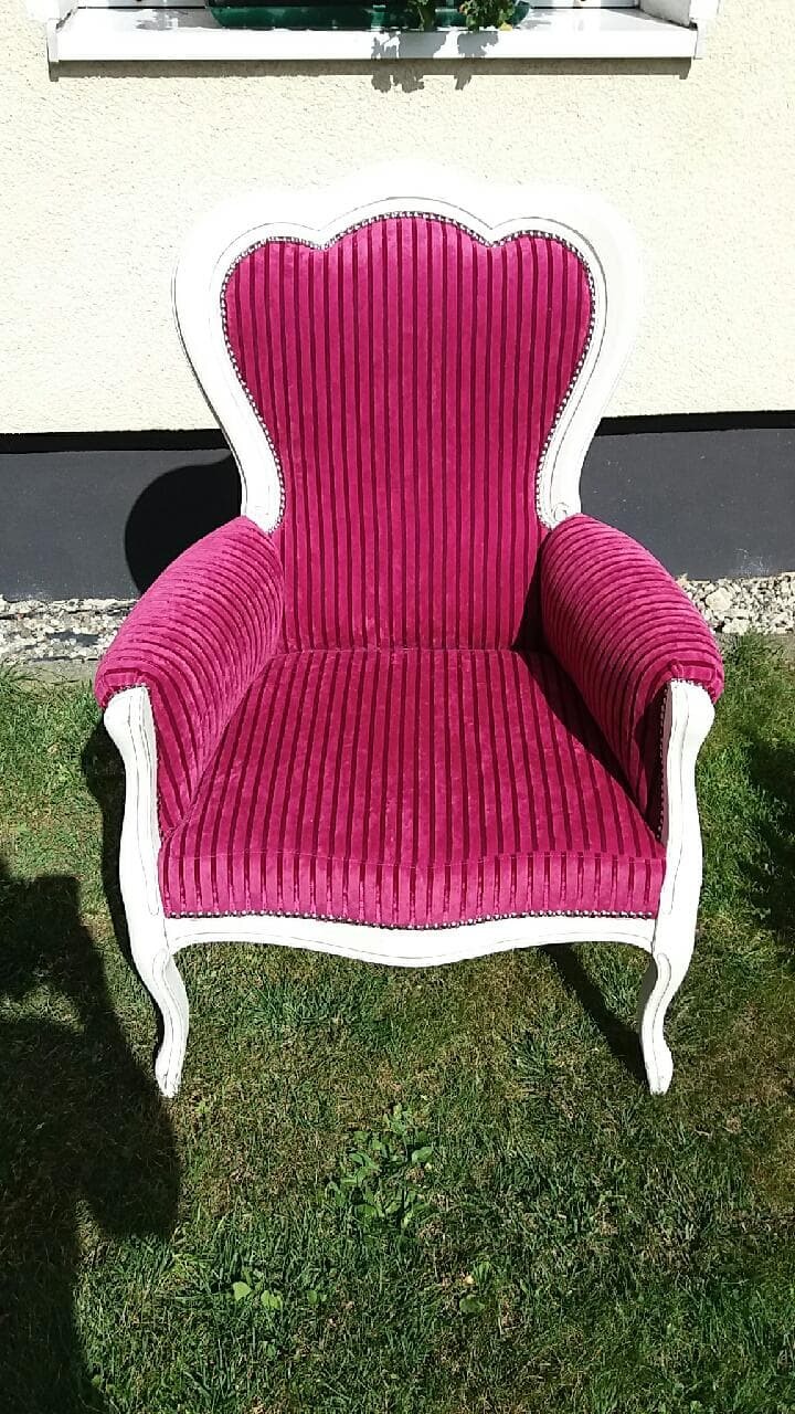 Fotel w stylu Ludwika XVI z różową, prążkowaną tapicerką i białą ramą, stojący na trawie przed jasną ścianą budynku.
