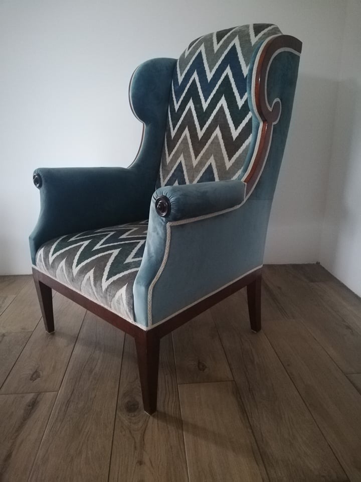 Błękitny fotel uszak z geometrycznym wzorem w zygzaki na oparciu i siedzisku, drewniane nogi, widoczny na tle białej ściany i drewnianej podłogi.