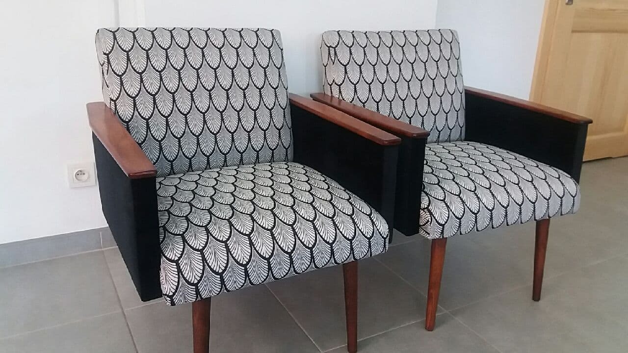 Dwa odnowione fotele z drewnianymi podłokietnikami i tapicerką w geometryczny wzór piór na szarym tle podłogi i białej ściany z gniazdkiem elektrycznym.