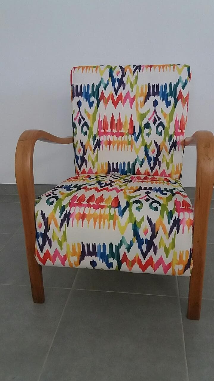 Odrestaurowany fotel z drewnianymi podłokietnikami, obity tkaniną w kolorowe, geometryczne wzory na białym tle.