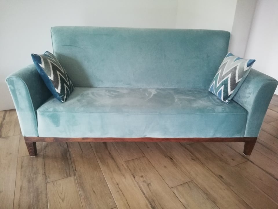 Turkusowa sofa z weluru z drewnianymi nogami i dwoma poduszkami w zygzaki na drewnianej podłodze.
