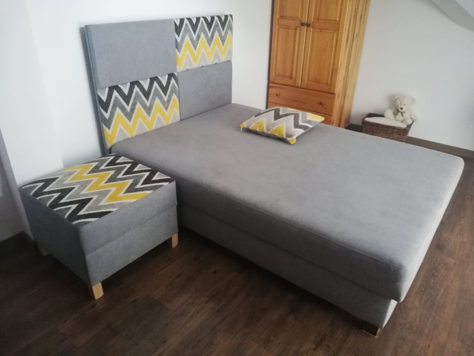 Szare łóżko z tapicerowanym zagłówkiem w zygzaki (żółty, szary, czarny) i pufą, drewniana szafa w tle, na łóżku poduszka w tym samym wzorze