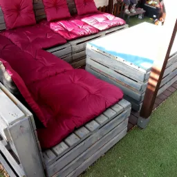 Meble ogrodowe z palet pomalowanych na szaro, z bordowymi poduchami, ustawione na sztucznej trawie.