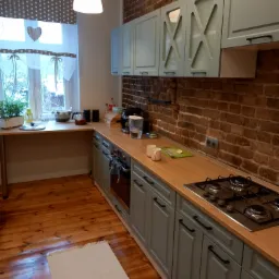 Kuchnia z szarymi szafkami, drewnianym blatem i ceglaną ścianą w tle, widok na okno z zasłonami w kropki i sercem.