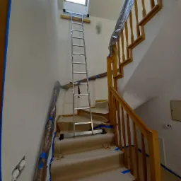 Schody zabezpieczone folią i taśmą malarską przed malowaniem ścian; aluminiowa drabina oparta o ścianę pod oknem dachowym.