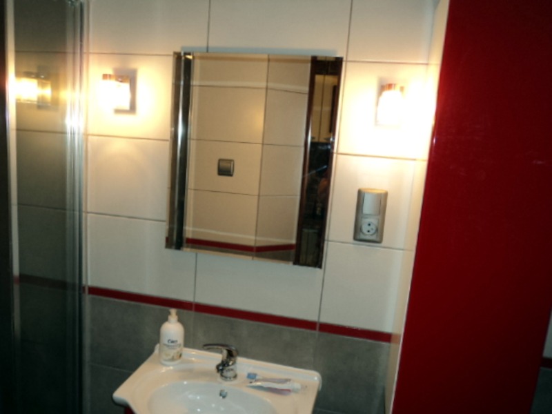 Wykończona łazienka z białymi płytkami, czerwonym akcentem, lustrem w metalowej ramie, umywalką z baterią, dozownikiem mydła i oświetleniem punktowym.