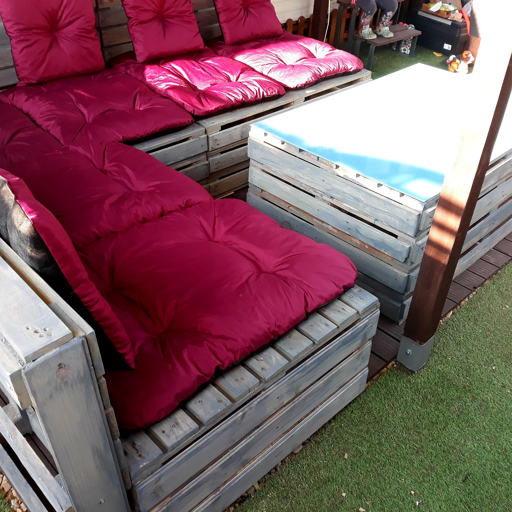 Meble ogrodowe z palet pomalowanych na szaro, z bordowymi poduchami, ustawione na sztucznej trawie.