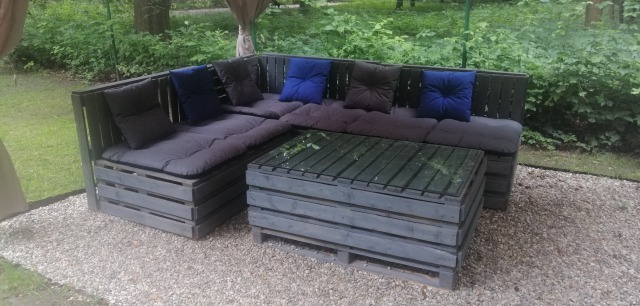 Drewniany narożnik ogrodowy z palet pomalowanych na szaro, z szarymi poduchami i granatowymi poduszkami dekoracyjnymi, stolik kawowy z palet, na żwirowym podłożu, w tle zieleń.