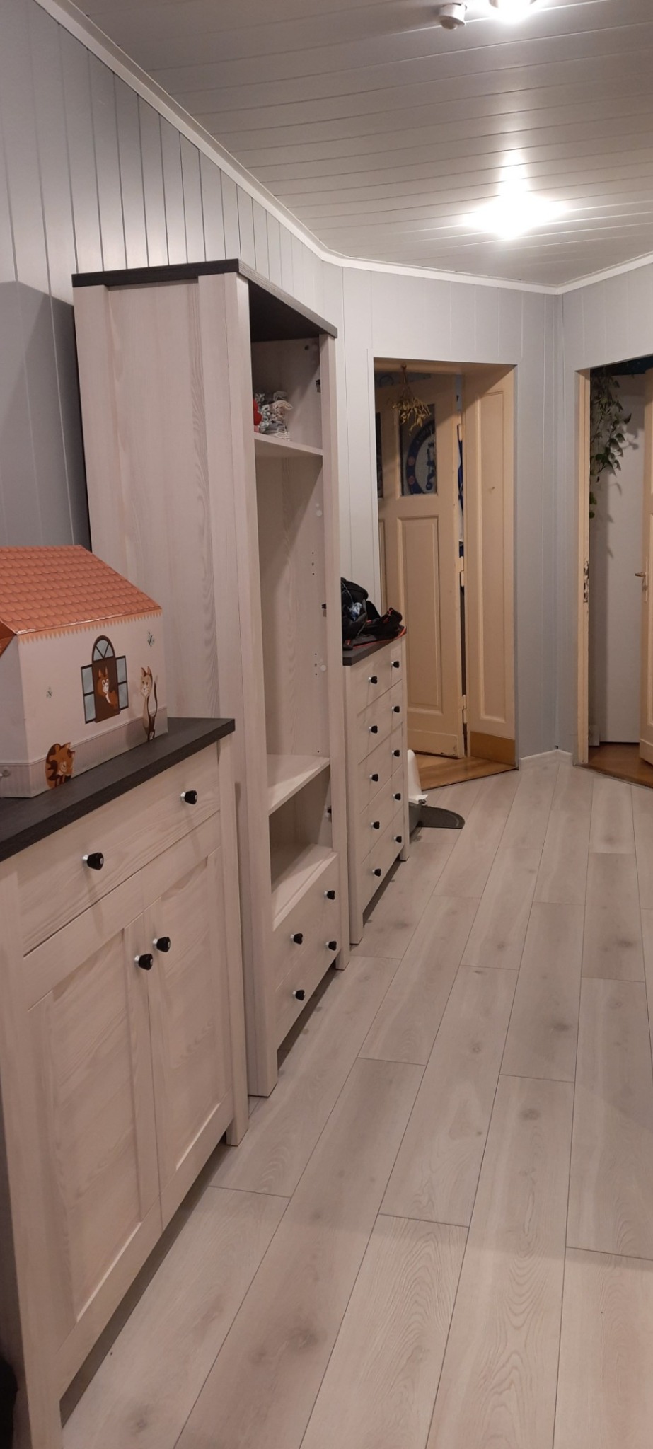 Półki i szafki z jasnego drewna z czarnymi blatami w pokoju z panelami podłogowymi i boazerią ścienną, widok na otwarte drzwi.