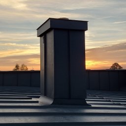 WRÓBELEK SPÓŁKA ZOO Piotr Wróbel - Szary, metalowy komin wentylacyjny na dachu z blachy na rąbek stojący, sfotografowany o wschodzie słońca.