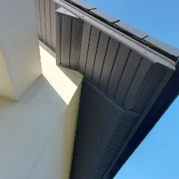 Nowoczesne zadaszenie z blachy trapezowej w kolorze grafitowym, widok z dołu na tle błękitnego nieba, fragment elewacji budynku w jasnym odcieniu.