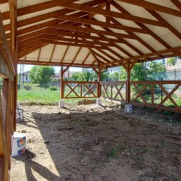 WRÓBELEK SPÓŁKA ZOO Piotr Wróbel - Wnętrze drewnianej altany z białym dachem w trakcie budowy, widoczne puste wiadra po farbie i częściowo ukończone ogrodzenie z krzyżowymi belkami.
