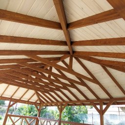 WRÓBELEK SPÓŁKA ZOO Piotr Wróbel - Drewniana konstrukcja z jasnym, panelowym dachem, widok od dołu, ukazujący skomplikowany układ belek i zadaszenie, z fragmentami otoczenia na zewnątrz.