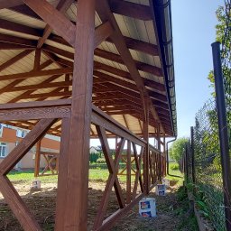 WRÓBELEK SPÓŁKA ZOO Piotr Wróbel - Konstrukcja z ciemnego drewna z jasnym dachem, osadzona na betonowych fundamentach w trakcie budowy, widoczne puszki po farbie i ogrodzenie z siatki.