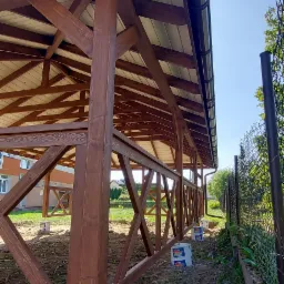 Konstrukcja z ciemnego drewna z jasnym dachem, osadzona na betonowych fundamentach w trakcie budowy, widoczne puszki po farbie i ogrodzenie z siatki.