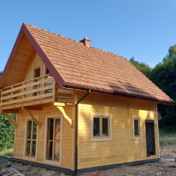 WRÓBELEK SPÓŁKA ZOO Piotr Wróbel - Surowy drewniany dom z balkonem i czerwoną dachówką na tle drzew i błękitnego nieba, widać elementy budowlane na ziemi.