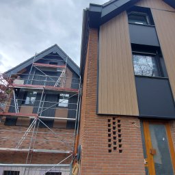 WRÓBELEK SPÓŁKA ZOO Piotr Wróbel - Montaż elewacji domu: widok na dwa budynki z rusztowaniem, jeden z brązową elewacją imitującą drewno i czarnymi panelami, drugi z czarną elewacją i podobnymi panelami. Na rusztowaniu widoczna osoba...