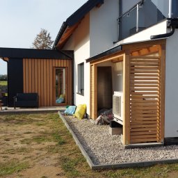 WRÓBELEK SPÓŁKA ZOO Piotr Wróbel - Nowoczesny dom z elementami drewna, widoczna drewniana osłona na klimatyzator z poziomymi listwami, oraz drewniane wykończenie ściany. Na zewnątrz widoczne meble ogrodowe i jacuzzi.