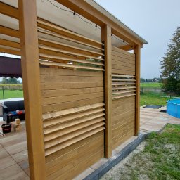 WRÓBELEK SPÓŁKA ZOO Piotr Wróbel - Drewniana konstrukcja z lamelami, częściowo zadaszona, oświetlona żarówkami, na tarasie z kostki, z widokiem na ogród i basen.