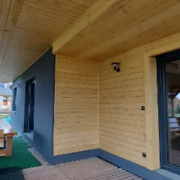 Zadaszony taras z drewnianą podłogą i elewacją, fragment ściany pomalowany na ciemny kolor, widoczne okno i drzwi przesuwne, lampa zewnętrzna, w tle domy jednorodzinne.