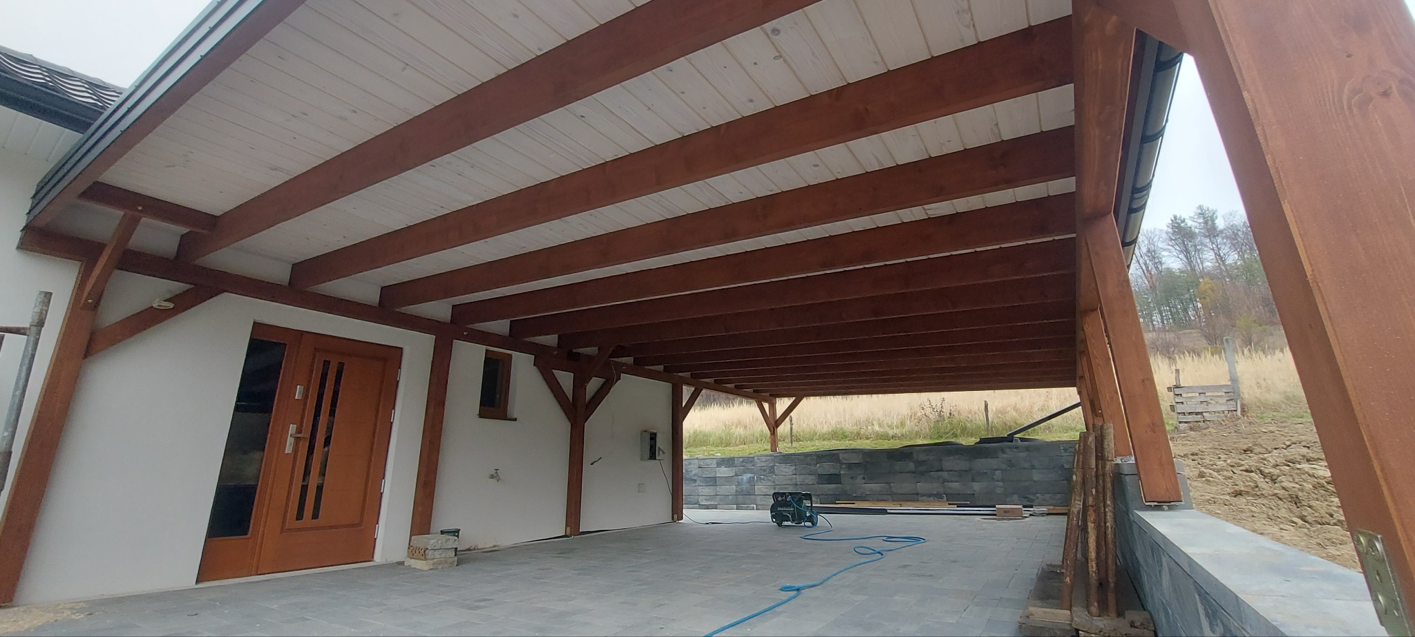 Zadaszenie tarasu z drewnianą konstrukcją wspartą na słupach, widoczne białe deski sufitowe i brązowe belki, na podłodze szara kostka brukowa, w tle mur z szarych bloczków.