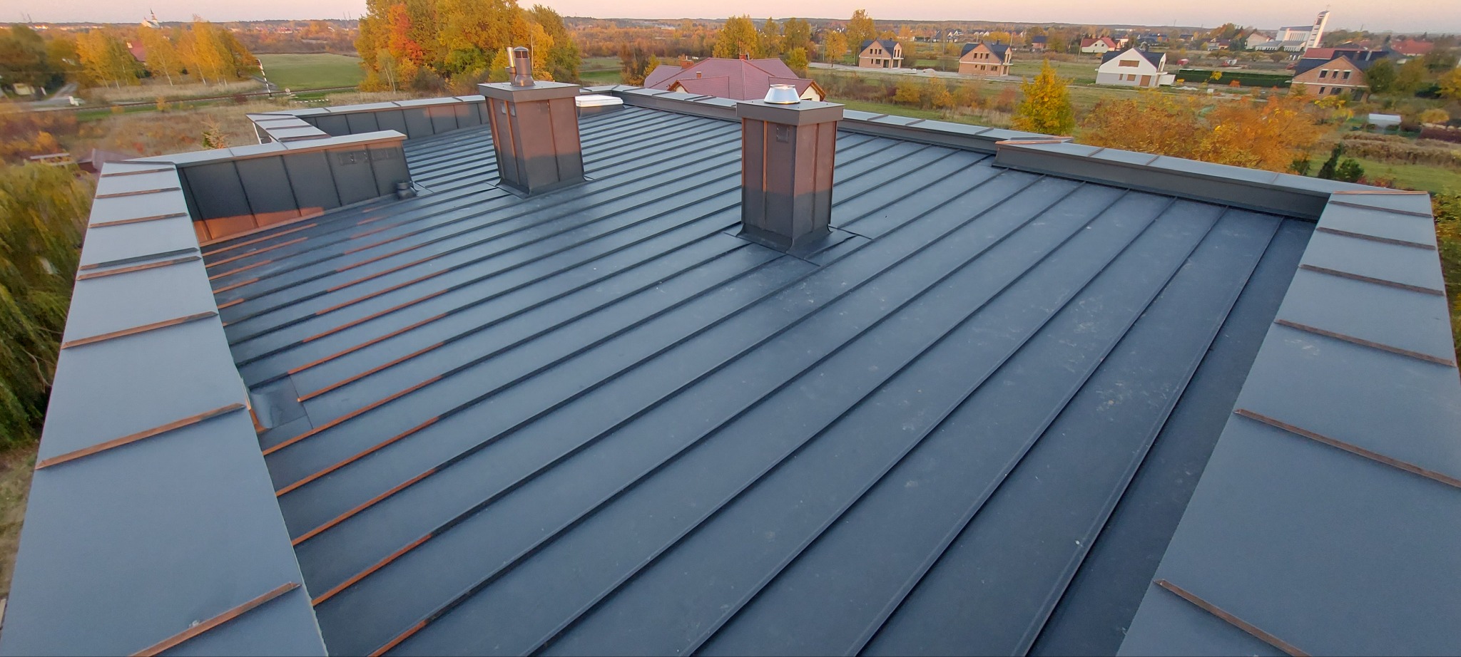 Nowoczesny, szary dach z blachy na domu jednorodzinnym, widok z góry, dwa kominy z metalowym wykończeniem, w tle zabudowa mieszkalna i jesienne drzewa.