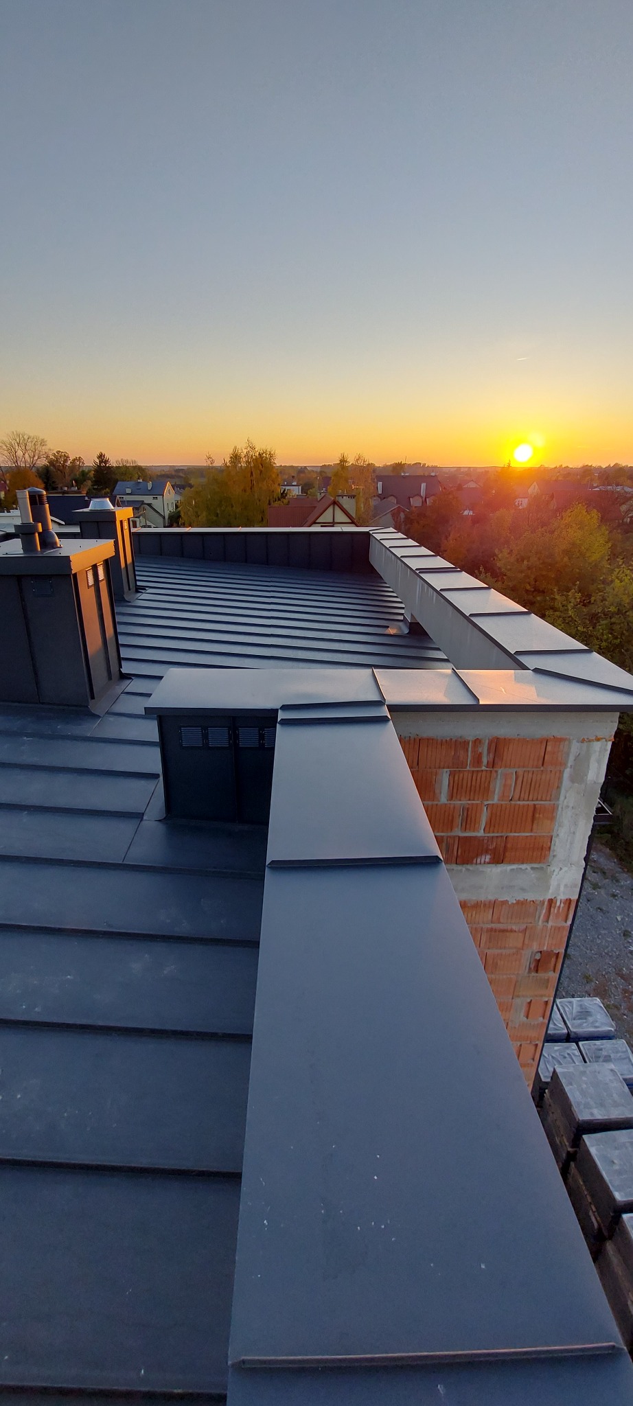 Nowoczesny dach z blachy na domu jednorodzinnym, widok z góry na tle zachodzącego słońca, komin w trakcie budowy.