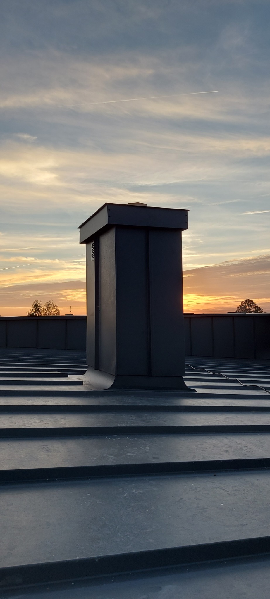 Szary, metalowy komin wentylacyjny na dachu z blachy na rąbek stojący, sfotografowany o wschodzie słońca.