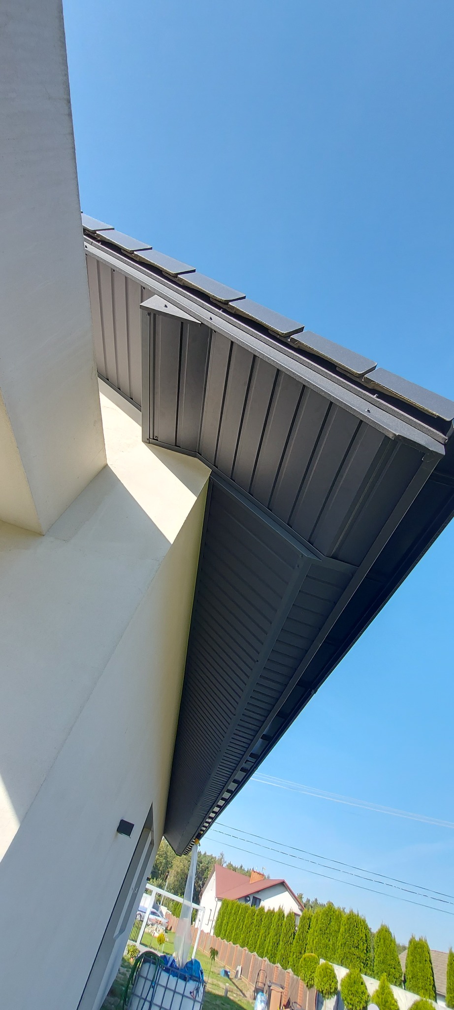 Nowoczesne zadaszenie z blachy trapezowej w kolorze grafitowym, widok z dołu na tle błękitnego nieba, fragment elewacji budynku w jasnym odcieniu.