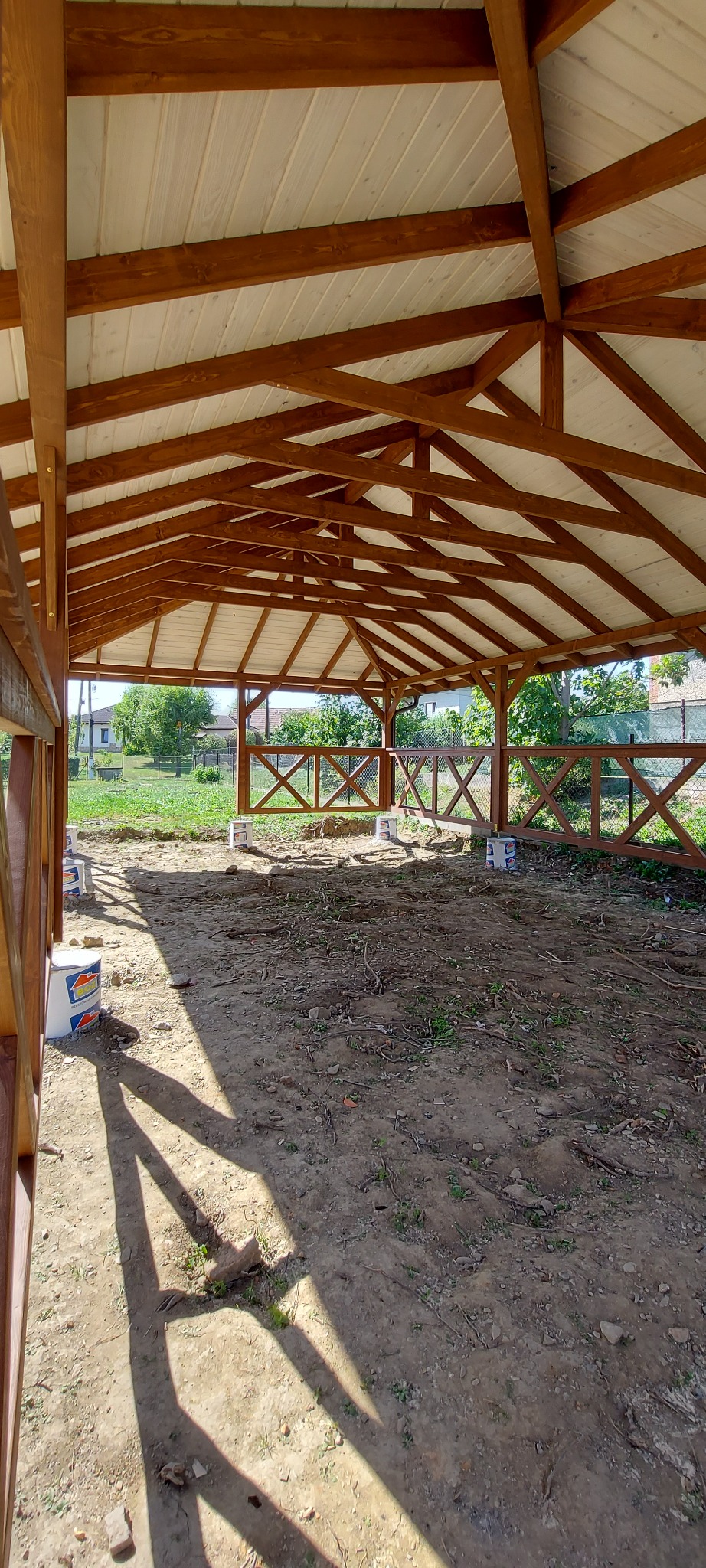 Wnętrze drewnianej altany z białym dachem w trakcie budowy, widoczne puste wiadra po farbie i częściowo ukończone ogrodzenie z krzyżowymi belkami.