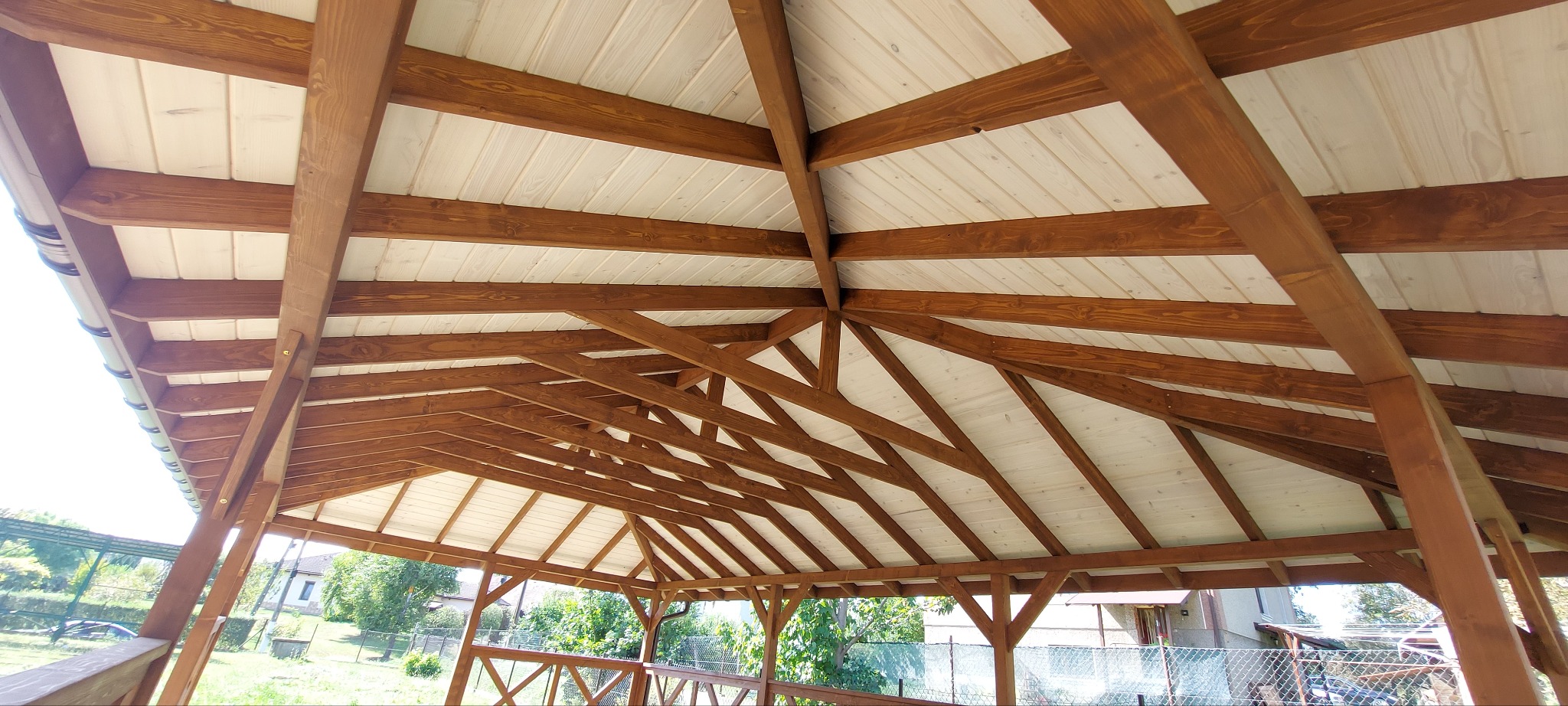 Drewniana konstrukcja z jasnym, panelowym dachem, widok od dołu, ukazujący skomplikowany układ belek i zadaszenie, z fragmentami otoczenia na zewnątrz.