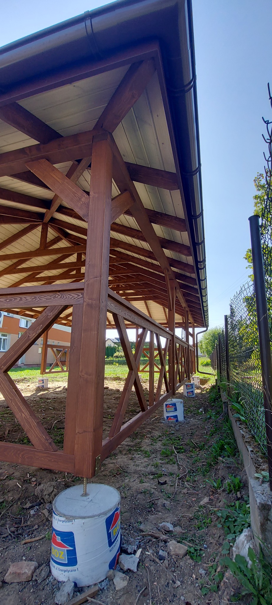 Konstrukcja z ciemnego drewna z jasnym dachem, osadzona na betonowych fundamentach w trakcie budowy, widoczne puszki po farbie i ogrodzenie z siatki.