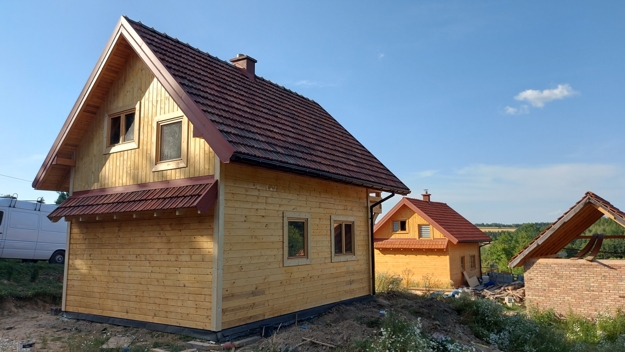 Nowo wybudowany, drewniany dom z czerwonym dachem, widok z boku, obok drugi dom w podobnym stylu oraz fragment murowanej konstrukcji.