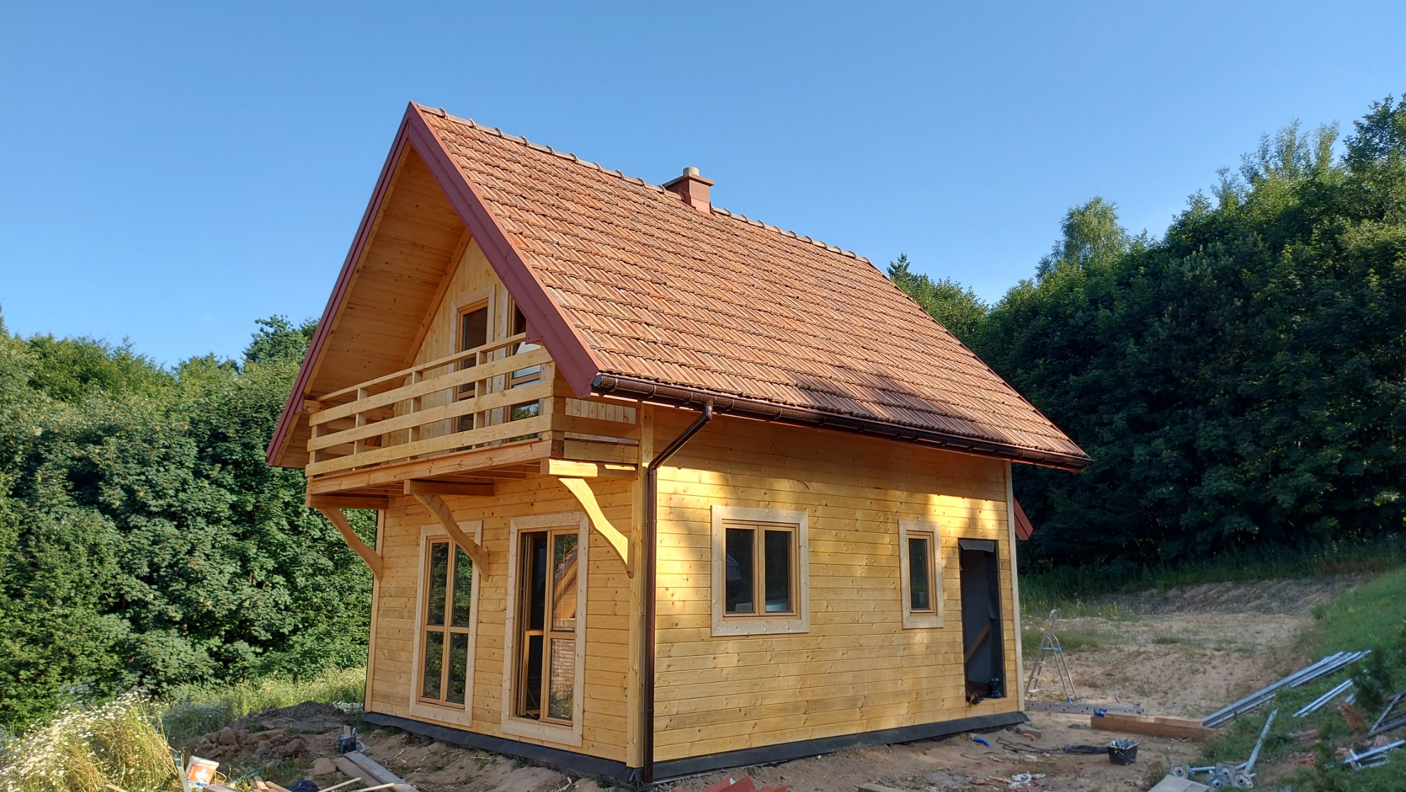 Surowy drewniany dom z balkonem i czerwoną dachówką na tle drzew i błękitnego nieba, widać elementy budowlane na ziemi.