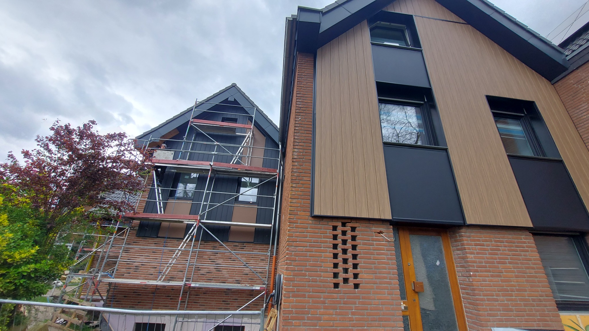 Montaż elewacji domu: widok na dwa budynki z rusztowaniem, jeden z brązową elewacją imitującą drewno i czarnymi panelami, drugi z czarną elewacją i podobnymi panelami. Na rusztowaniu widoczna osoba...