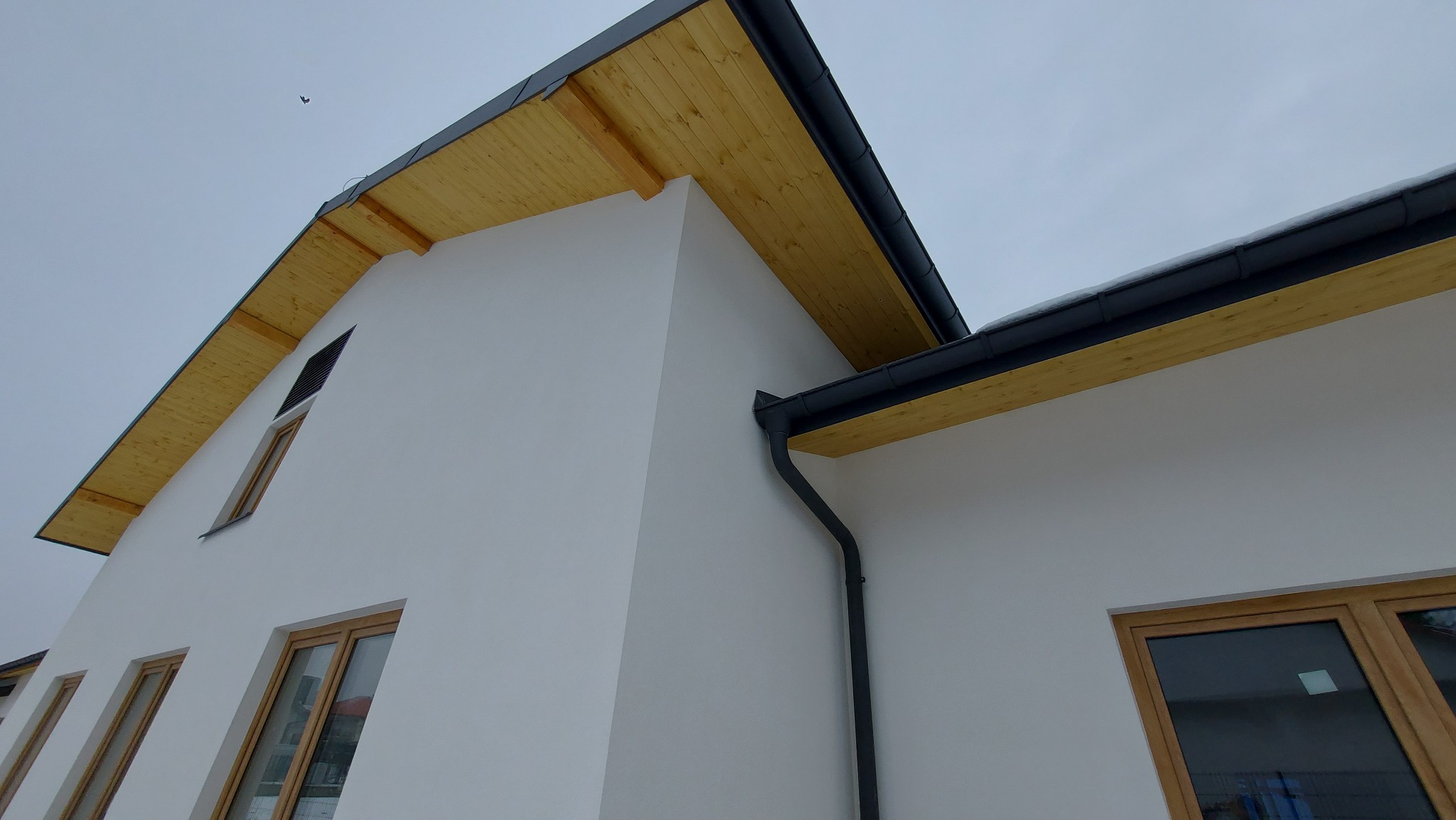 Fragment białego budynku z drewnianym podbitkiem dachowym i czarnymi rynnami, widok z dołu, z widocznym fragmentem okna i niebem w tle.