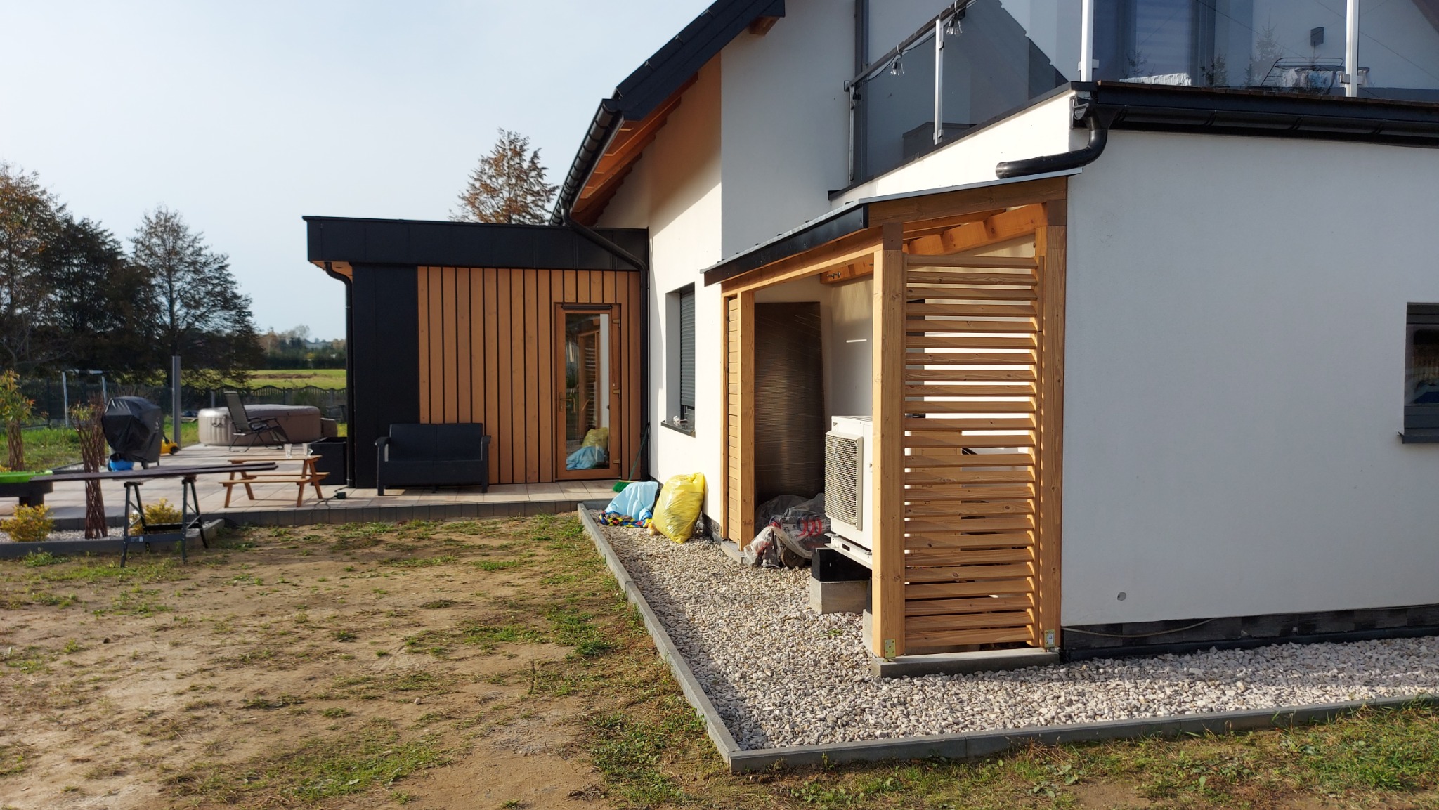 Nowoczesny dom z elementami drewna, widoczna drewniana osłona na klimatyzator z poziomymi listwami, oraz drewniane wykończenie ściany. Na zewnątrz widoczne meble ogrodowe i jacuzzi.