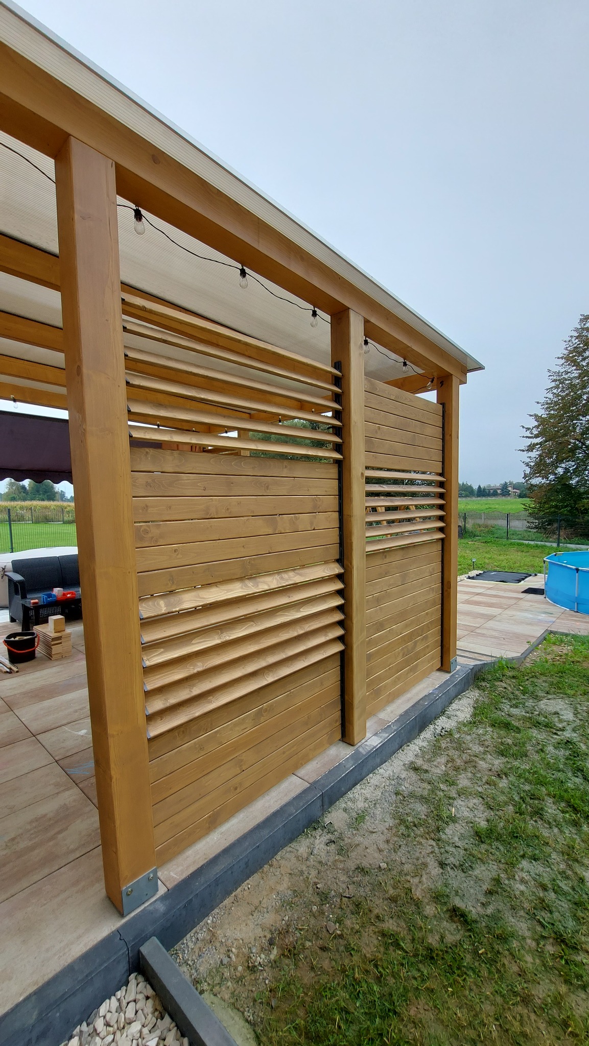 Drewniana konstrukcja z lamelami, częściowo zadaszona, oświetlona żarówkami, na tarasie z kostki, z widokiem na ogród i basen.