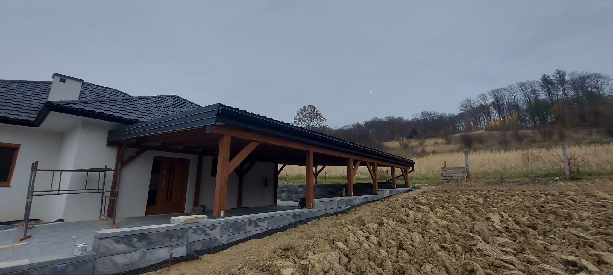 Zadaszenie tarasu z drewnianą konstrukcją w Pustkowie, widok z boku na tle zaoranego pola i wzgórza z drzewami, obok domu z ciemną dachówką.