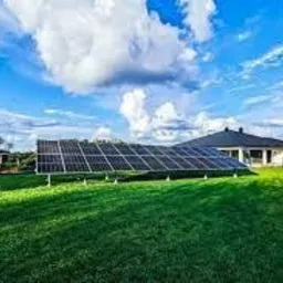 Panel słoneczny na zielonym trawniku przed nowoczesnym domem jednorodzinnym z ciemnym dachem, błękitne niebo z białymi chmurami w tle.