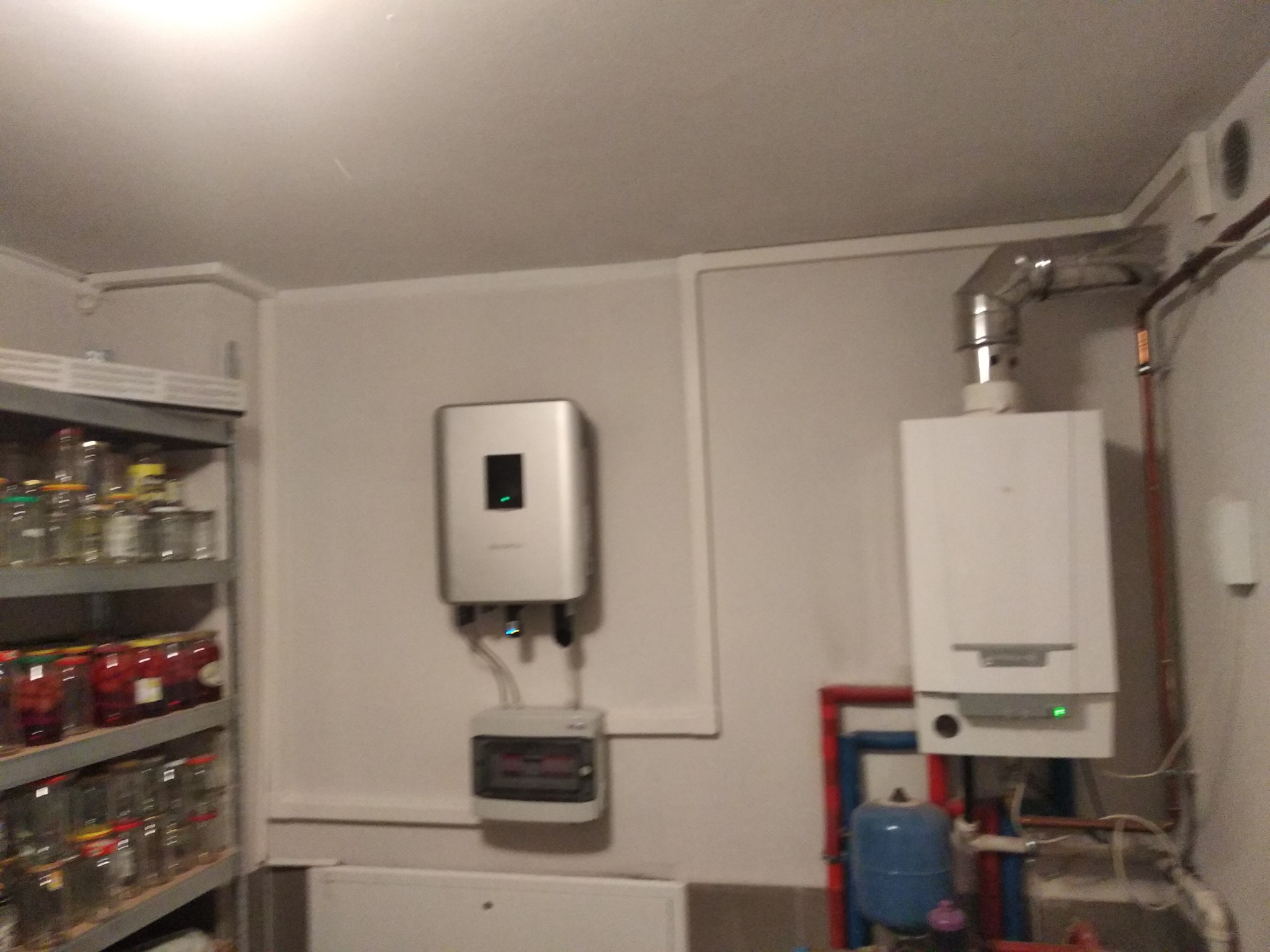 Wnętrze pomieszczenia z zamontowanym srebrnym inwerterem fotowoltaicznym nad szarą skrzynką bezpieczników, obok biały piec gazowy z instalacją hydrauliczną i półka z przetworami w słoikach.