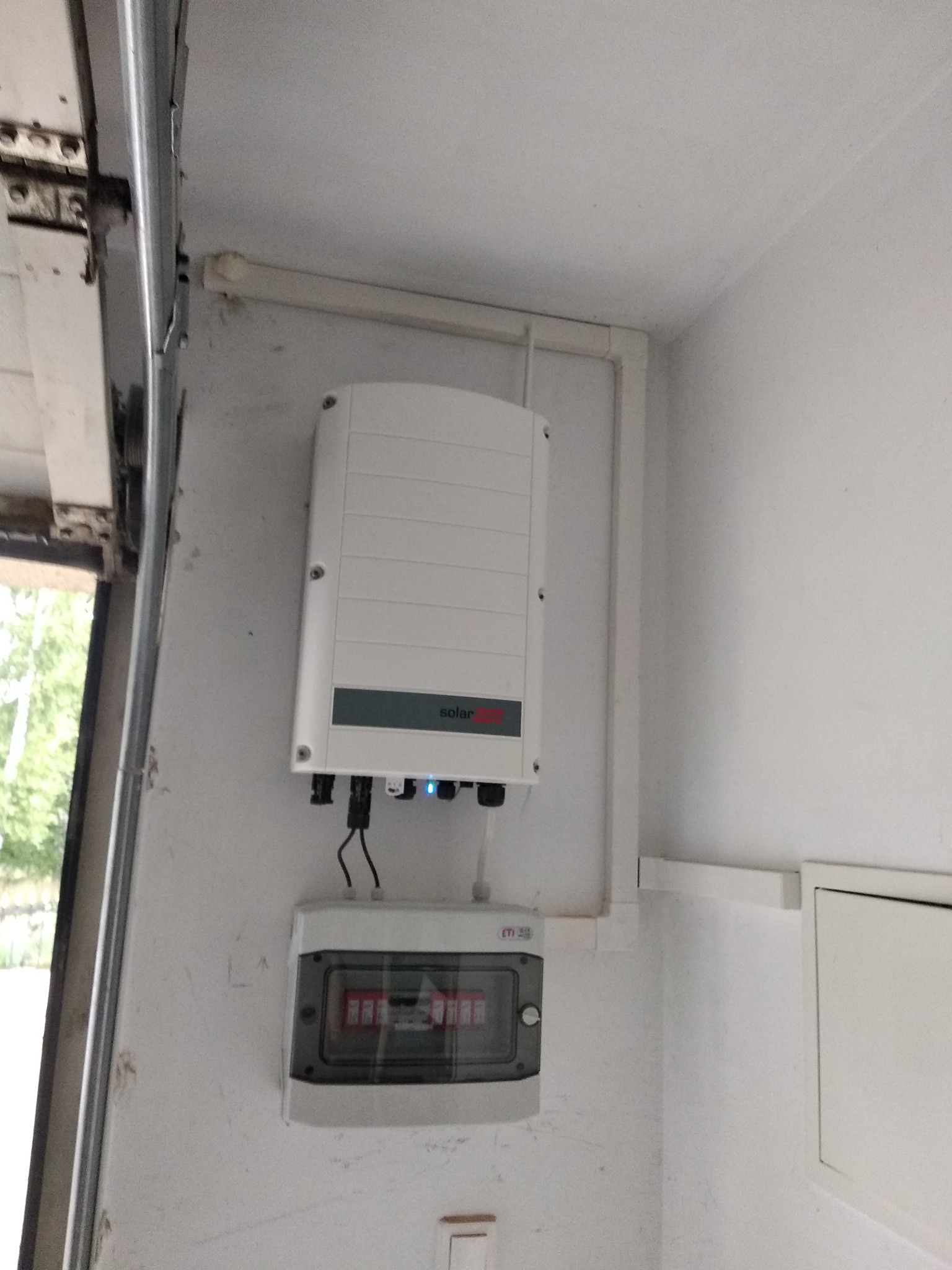 Biały falownik z niebieskimi diodami LED zamontowany na ścianie obok szarej skrzynki bezpiecznikowej, widoczne białe rurki instalacyjne i fragment otwartych drzwi garażowych z widokiem na zieleń.