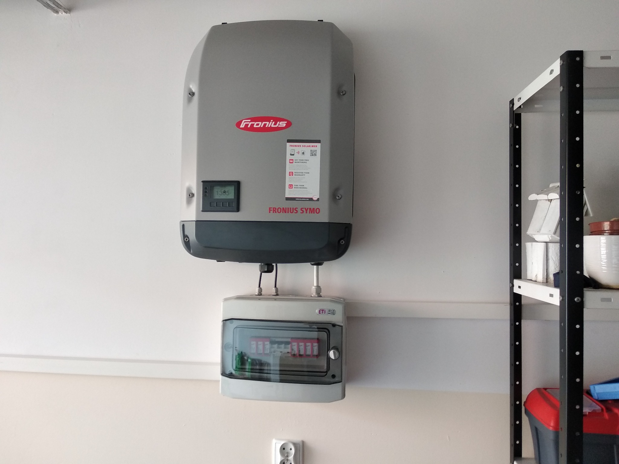 Zamontowany na ścianie szary falownik Fronius Symo z podłączoną skrzynką zabezpieczeń, widoczna instalacja elektryczna i fragment regału magazynowego.