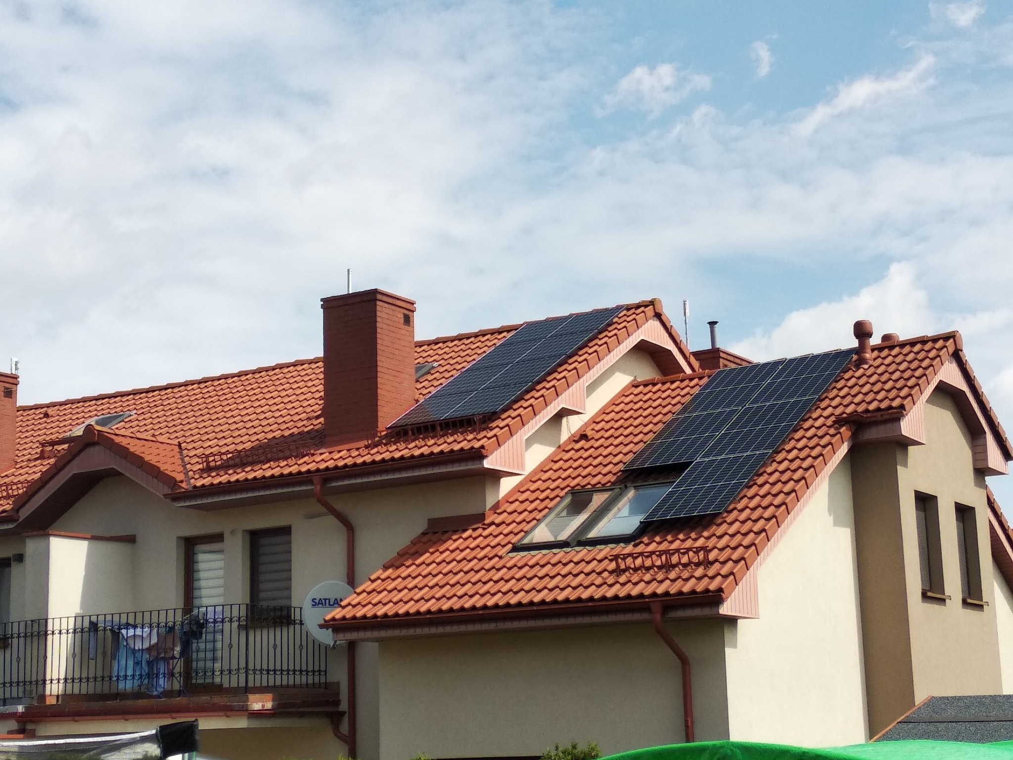 Panele słoneczne zamontowane na dachu domu z czerwoną dachówką, widoczne okna dachowe i komin, na balkonie suszące się pranie, antena satelitarna SATLAN na ścianie budynku.