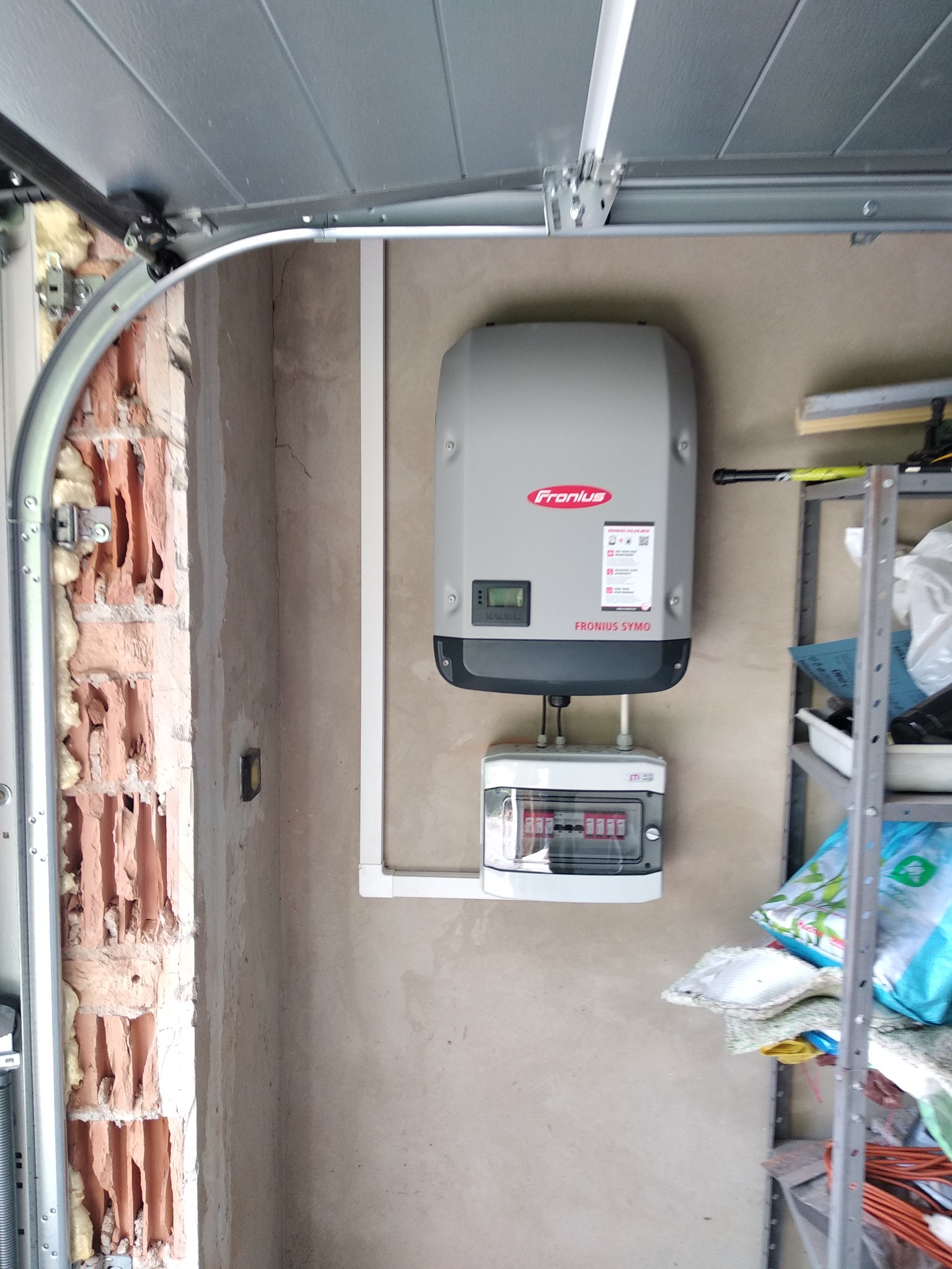 Szary falownik Fronius SYMO zamontowany na ścianie w garażu, nad skrzynką bezpiecznikową, widoczna boczna ściana z cegieł i fragment otwartej bramy garażowej.